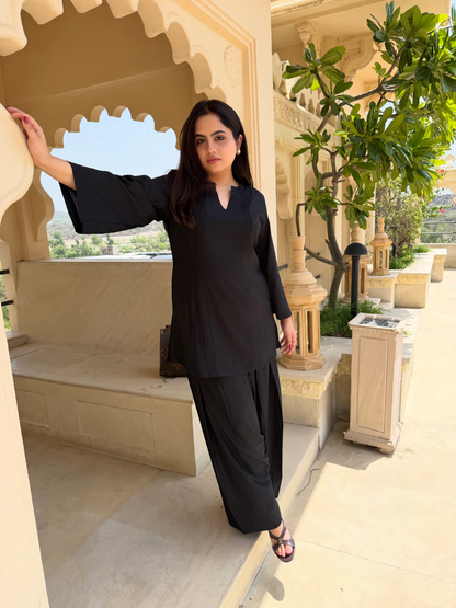 Woman in black farsi salwar suit  outfit standing in an ornate architectural setting with greenery.