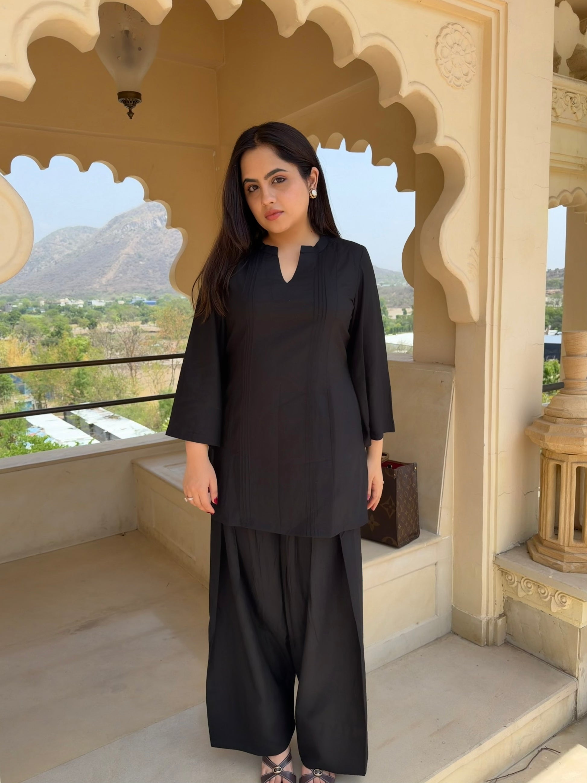 Woman in a black farsi salwar suit  outfit standing in an ornate architectural setting with mountains in the background