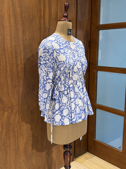 A blue and white floral print long-sleeve blouse displayed on a beige dress form mannequin, set against a wooden wall and glass-paneled door.
